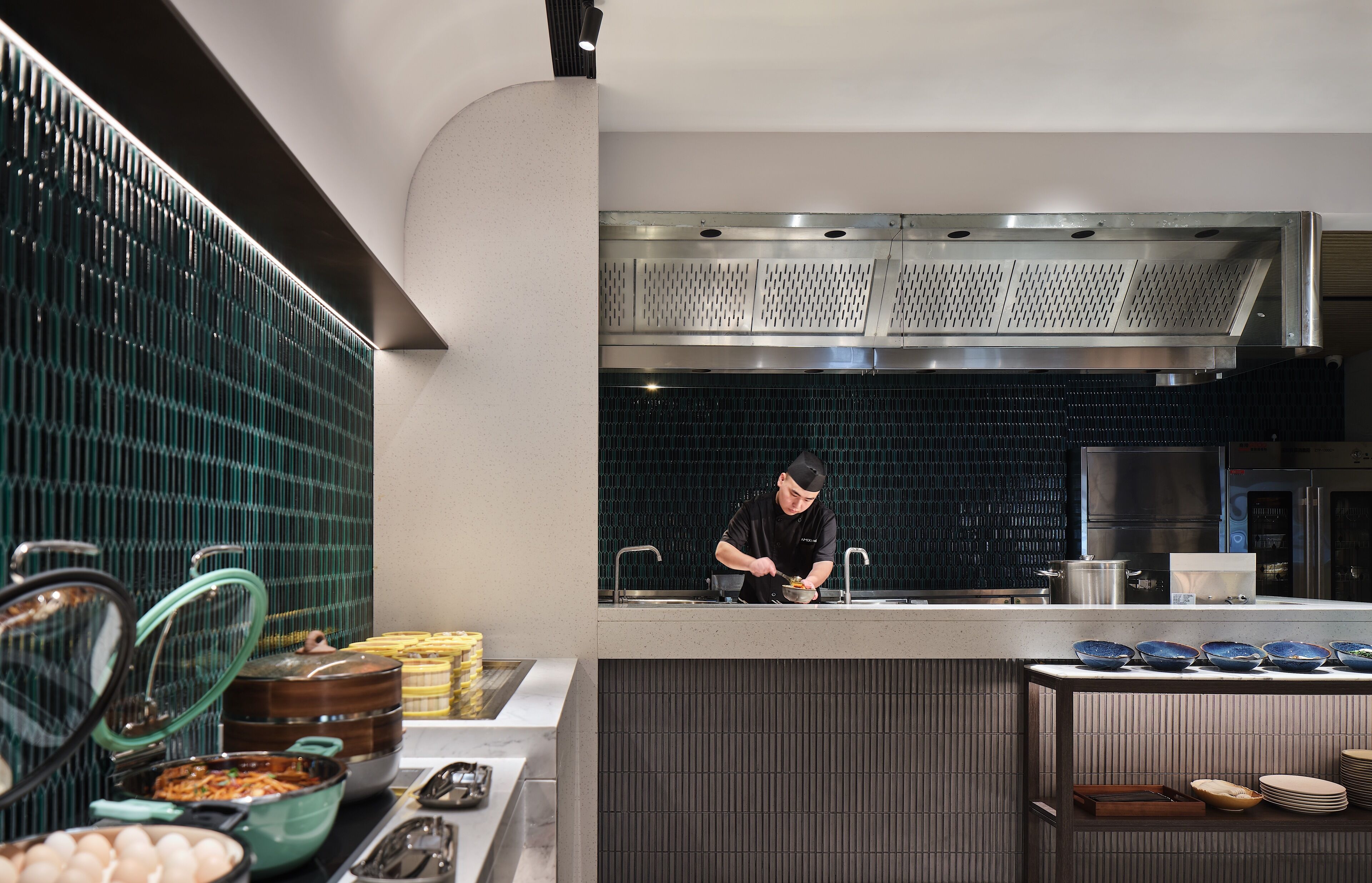 Une cuisine moderne où un chef prépare des plats, avec une variété d’ustensiles de cuisine et un buffet dressé avec des plats et des bols.