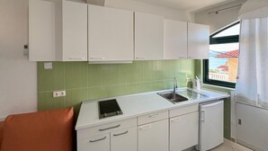 Een compacte keuken met witte kasten, een groene betegelde spatwand, een raam met uitzicht en een kleine oranje stoel.