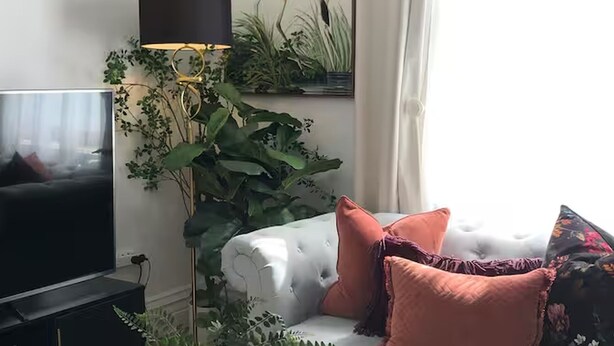 Un salon doté d’un grand téléviseur à écran plat, d’un canapé blanc agrémenté de plusieurs coussins et d’une grande plante surmontée d’une lampe.