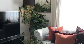 Un salon doté d’un grand téléviseur à écran plat, d’un canapé blanc agrémenté de plusieurs coussins et d’une grande plante surmontée d’une lampe.