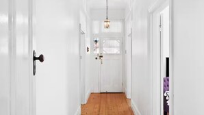 Un couloir blanc avec un plancher en bois, une porte dotée d’une poignée en laiton et un luminaire suspendu.