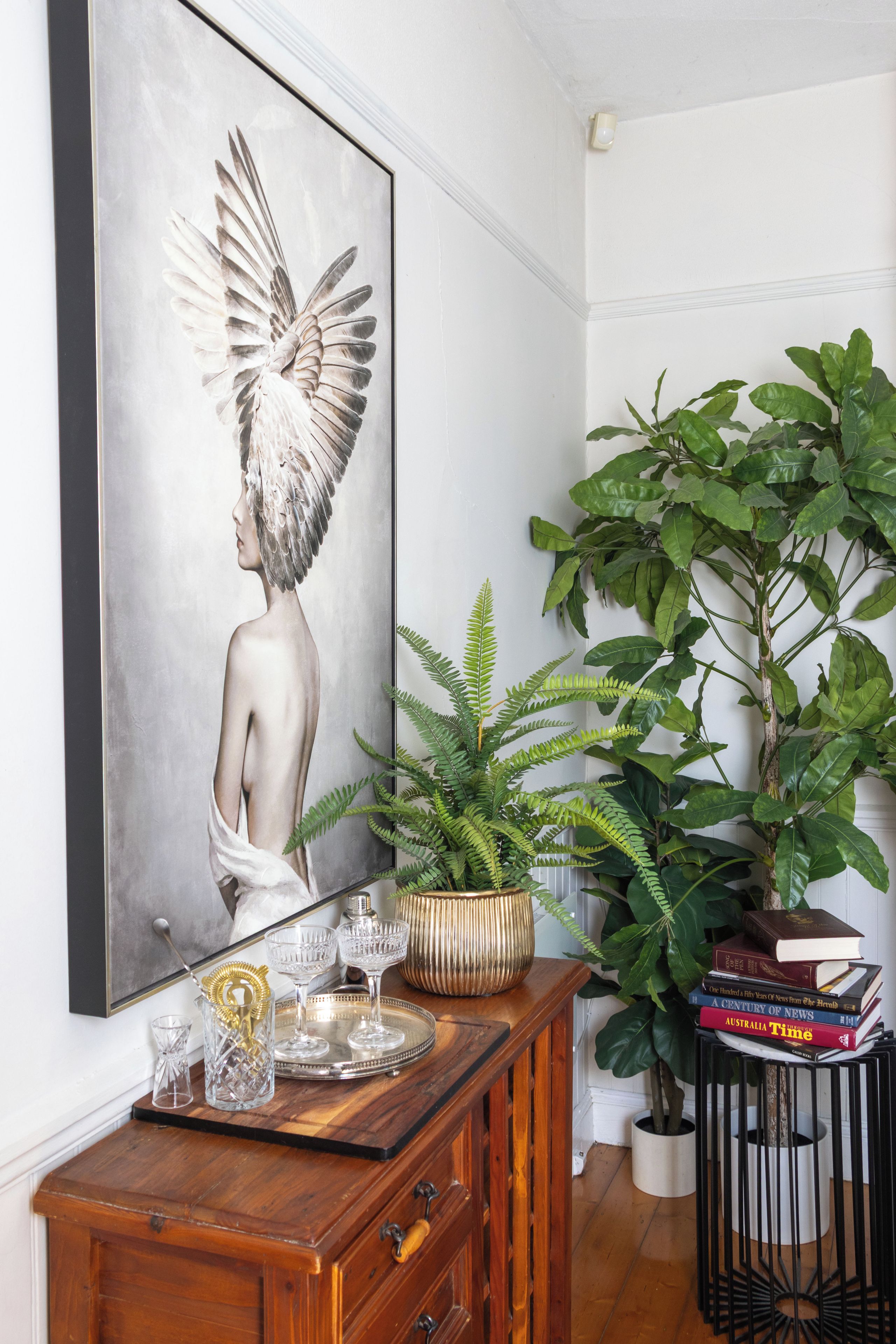 Una habitación con una obra de arte en un marco grande, un aparador de madera con cristalería, una planta en maceta y una pila de libros.