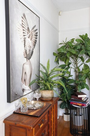 Una habitación con una obra de arte en un marco grande, un aparador de madera con cristalería, una planta en maceta y una pila de libros.