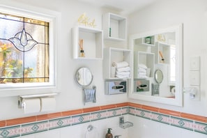 Une salle de bain avec une fenêtre, un miroir, des étagères contenant des articles de toilette et un panneau décoratif.