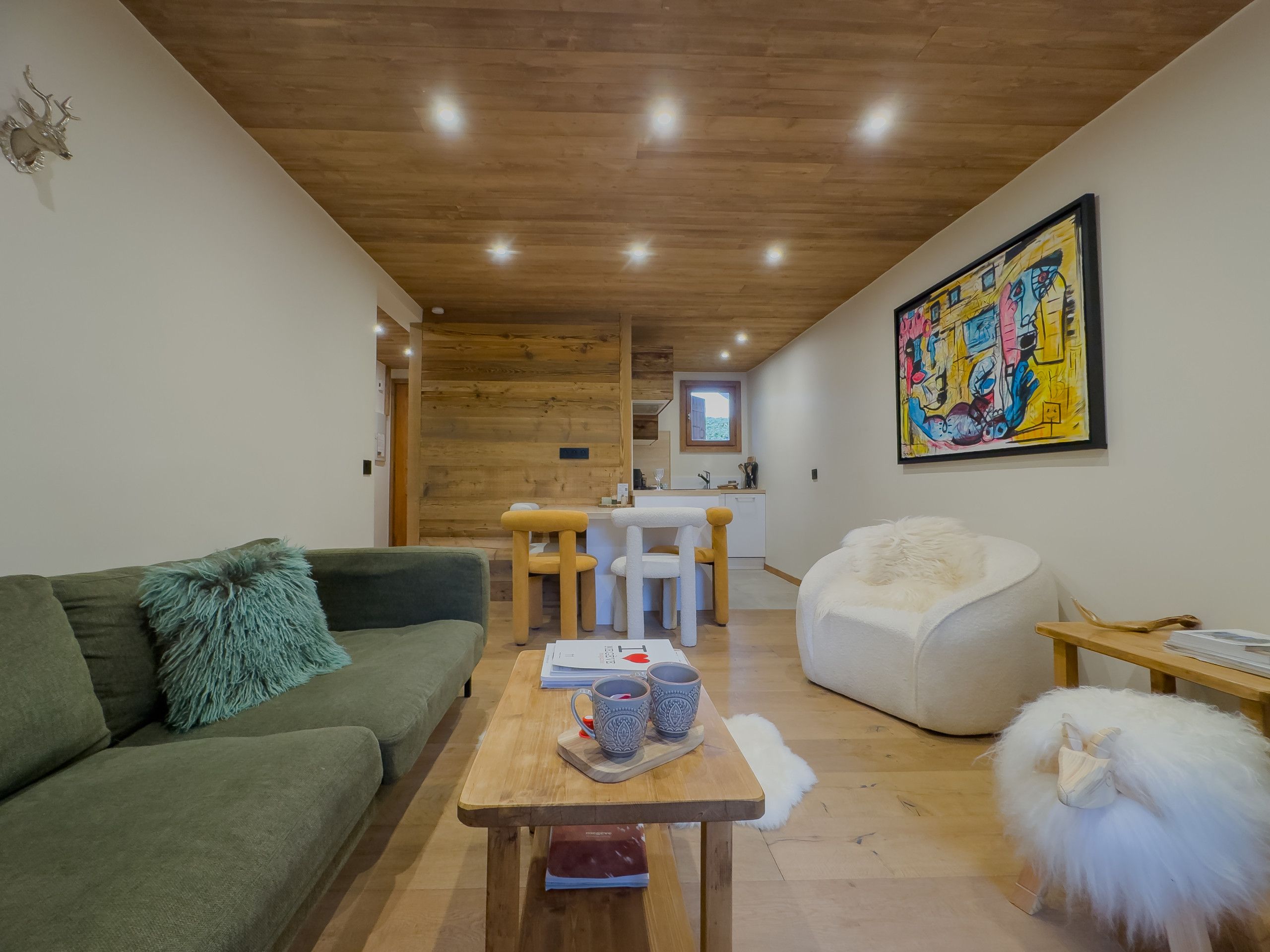 Ein modernes Wohnzimmer mit einem grünen Sofa, einem Couchtisch aus Holz mit Dekoration, einem weißen, flauschigen Sessel und einem farbenfrohen, abstrakten Gemälde an der Wand.