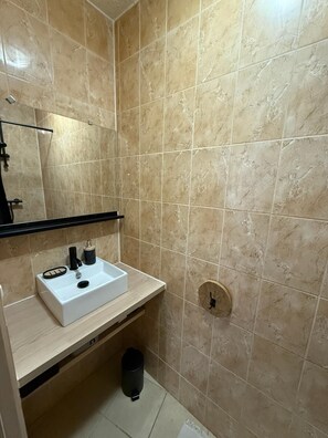 Un baño con baldosas de mármol color crema, un lavamanos blanco con dispensador de jabón negro, un espejo sobre el lavamanos y un cubo de basura negro debajo.