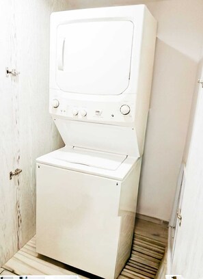 A compact, white washing machine in a small, enclosed space with a wooden floor and a white door. - Stylish Apt w/ In-Unit Laundry • Rooftop Pool (Playa del Carmen,)