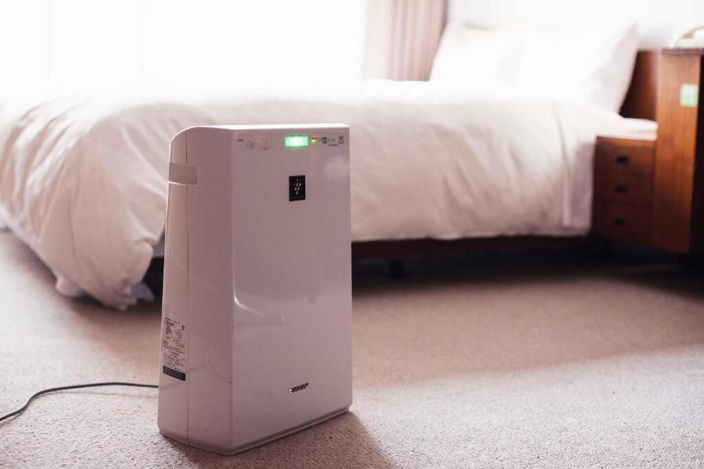 Calentador portátil blanco con panel de control y cable de alimentación, colocado sobre un suelo alfombrado en un dormitorio con cama y mesita de noche de madera.