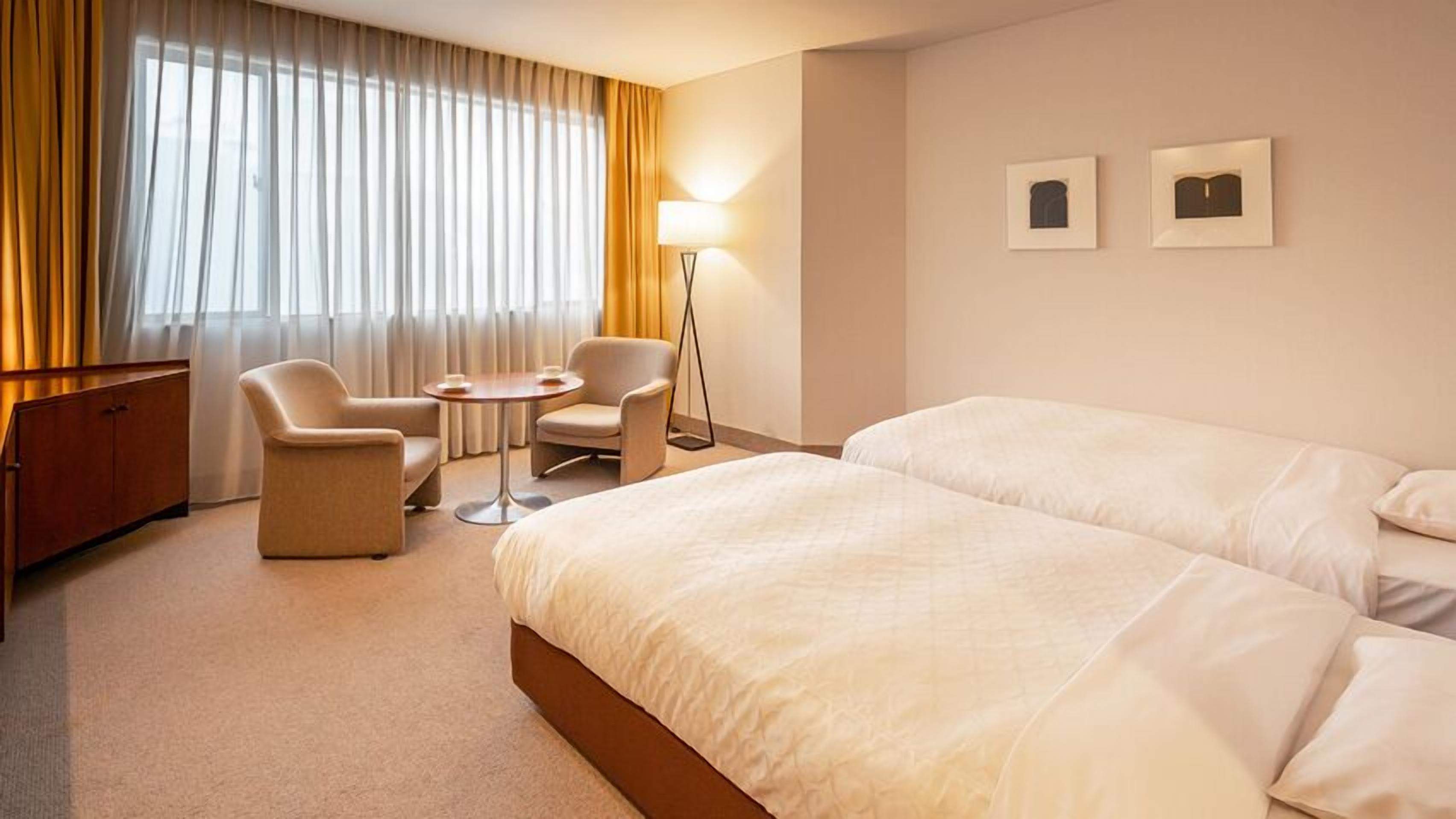 Habitació d'hotel amb dos llits, un escriptori, una cadira, una làmpada i dues fotografies enmarcades a la paret.