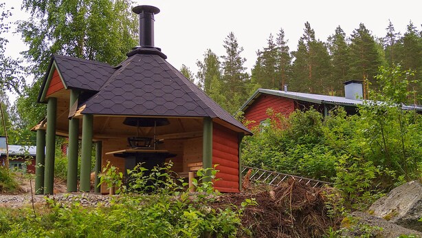Pieni puinen mökki, jossa on kuusikulmainen katto ja savupiippu, ja sen ympärillä on tiheää kasvillisuutta ja puita.