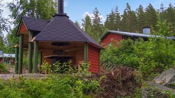 En liten stuga av trä med ett sexkantigt tak och en skorsten, omgiven av tät grönska och träd.