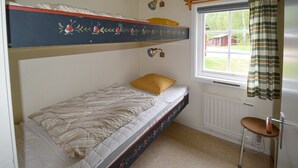 Et lille værelse med en enkeltseng og køjeseng, et vindue med udsigt til et hus, en radiator og en stol.