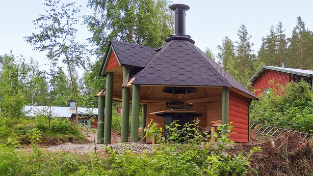 A small, hexagonal-roofed structure with a chimney, surrounded by greenery and a red building in the background. - Stunning home in Ekshärad with WiFi (Ekshärad)