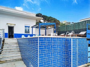 Ein Swimmingpool mit blauen Fliesen, ein überdachter Sitzbereich und eine Treppe, die zu einer Tür führt.