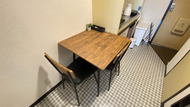 Een klein zithoekje met een houten tafel en twee stoelen. Op de tafel staat een kleine potplant, en naast de tafel bevindt zich een prullenbak. De vloer heeft een betegeld patroon.