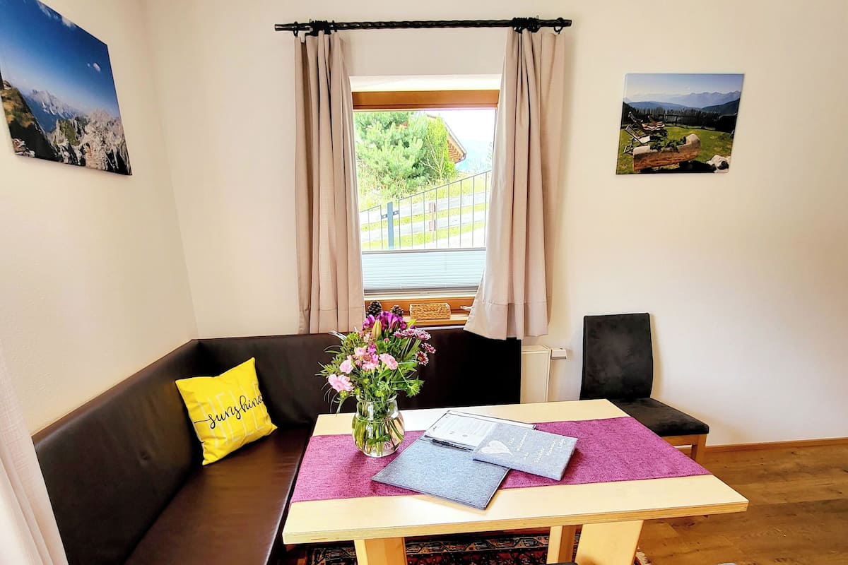 Een knus interieur met een houten vloer, een bruine bank met een geel kussen, een kleine tafel met een bloevas en twee stoelen.