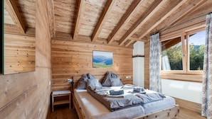 Een slaapkamer in een houten hutje, met een bed, kussens, een nachtkastje en een raam met gordijnen.