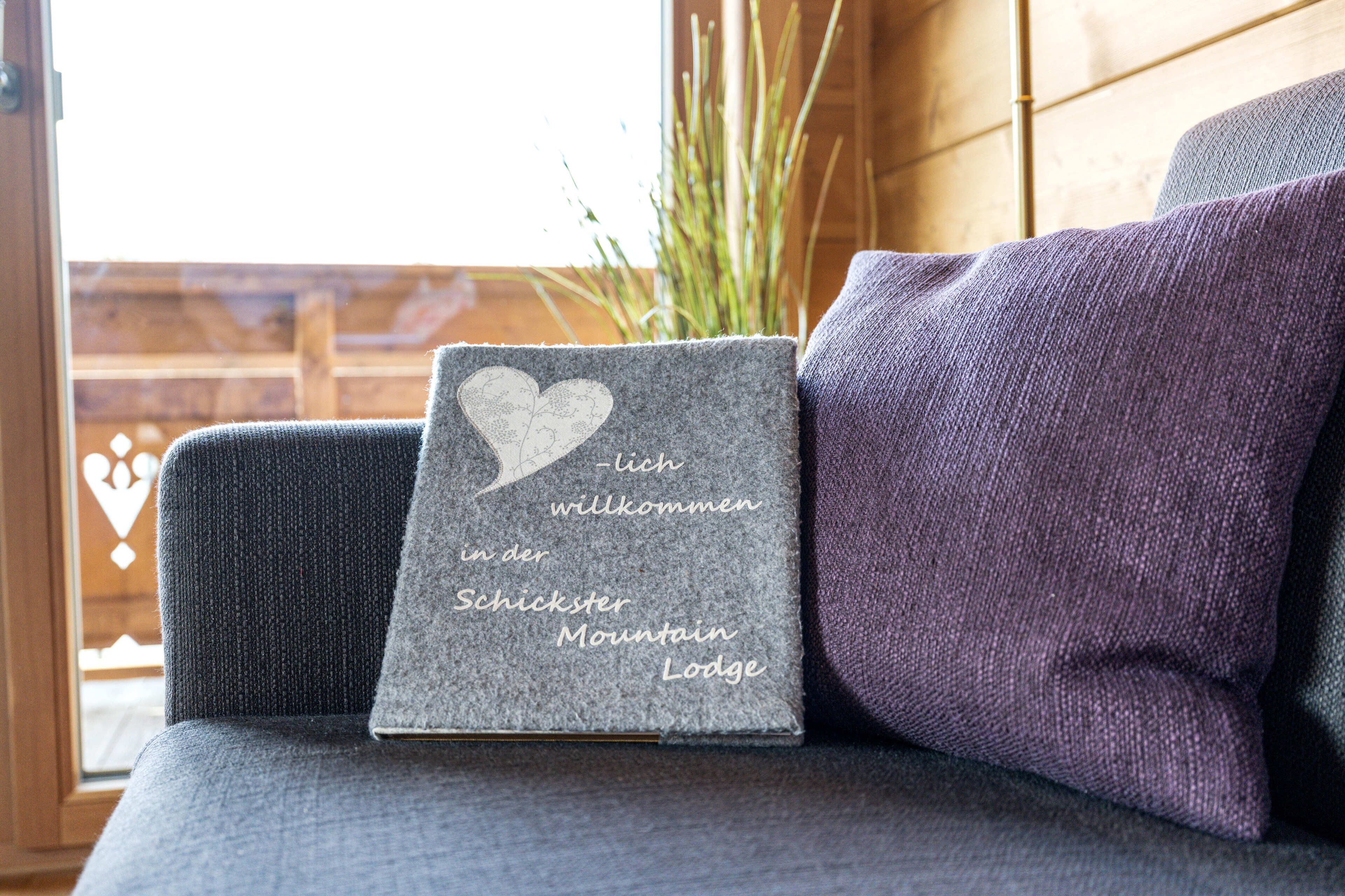 Un coussin gris orné d’une étiquette en forme de cœur sur laquelle on peut lire : « Ich willkommen in der Schickster Mountain Lodge », posé sur un canapé.