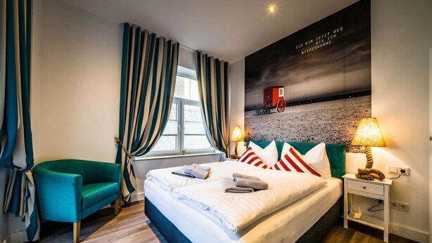 Quarto de hotel com uma cama grande, uma poltrona verde, uma mesinha de cabeceira com abajur e uma janela com cortinas.