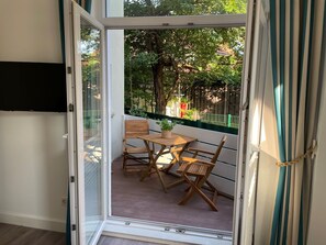 A balcony with a wooden table and chairs, a potted plant, and a view of trees and a fence.