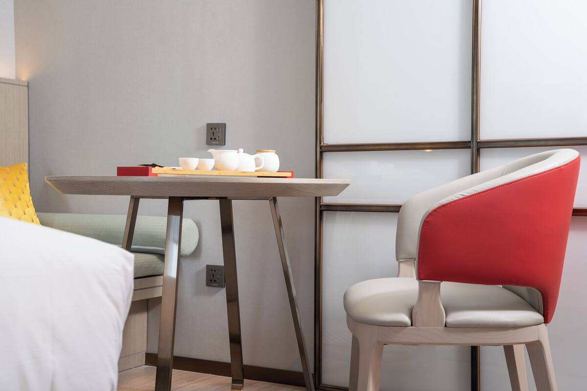 A hotel room with a bed, a table with a red book, a white chair, and a wall with a geometric pattern.
