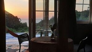 Ein Zimmer mit Meerblick bei Sonnenuntergang, ein Sessel, ein Tisch mit einer Vase und ein Fenster.