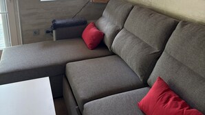 Ein modernes Interieur mit einem grauen Ecksofa, einem roten Kissen, einem Fenster mit Blick und einer weißen Wand.
