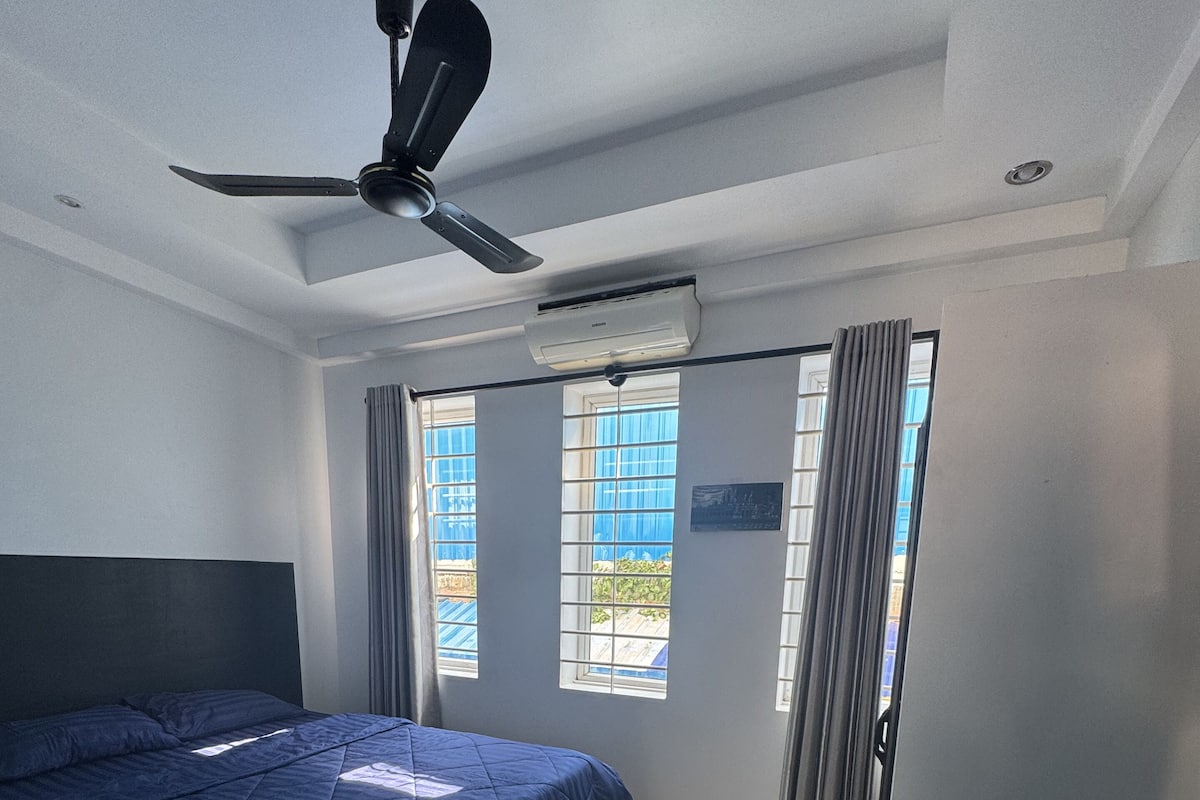 Habitación de hotel con cama, escritorio, silla, ventilador de techo y ventana con cortinas.