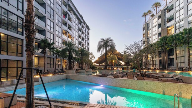 A hotel pool area with a large pool, lounge chairs, palm trees, and a thatched-roof structure. - Condo with Full Facilities C255 (Kamala)