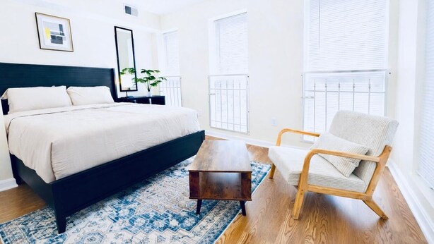 Ein modernes Schlafzimmer mit einem großen Bett, einem Holztisch, einem Sessel und einem gemusterten Teppich.