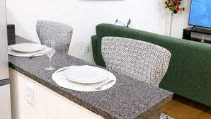 Ein kompakter Wohnraum mit Küchenzeile, einem grünen Sofa, einem kleinen Esstisch mit Tellern und einem Glas sowie einem Fernseher auf einem Schrank.