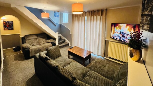 A cozy living room with a sofa, a coffee table, and a TV displaying a fireplace. A staircase with a railing and a painting on the wall are also visible. - Modern Comfortable Two Bedrooms House DL5 With Garden Parking and Free Wifi (Newton Aycliffe)