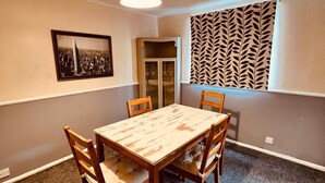 A dining area with a wooden table and chairs, a hanging light fixture, a framed cityscape picture, and a patterned window blind. - Modern Comfortable Two Bedrooms House DL5 With Garden Parking and Free Wifi (Newton Aycliffe)