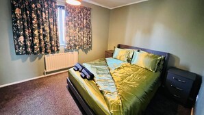 A bedroom with a bed, a nightstand, a dresser, a window with floral curtains, and a ceiling light. - Modern Comfortable Two Bedrooms House DL5 With Garden Parking and Free Wifi (Newton Aycliffe)