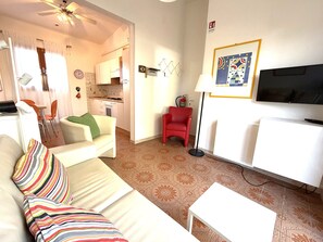 Una sala de estar con un sofá, una mesita, una silla roja, un televisor y una cocina al fondo.