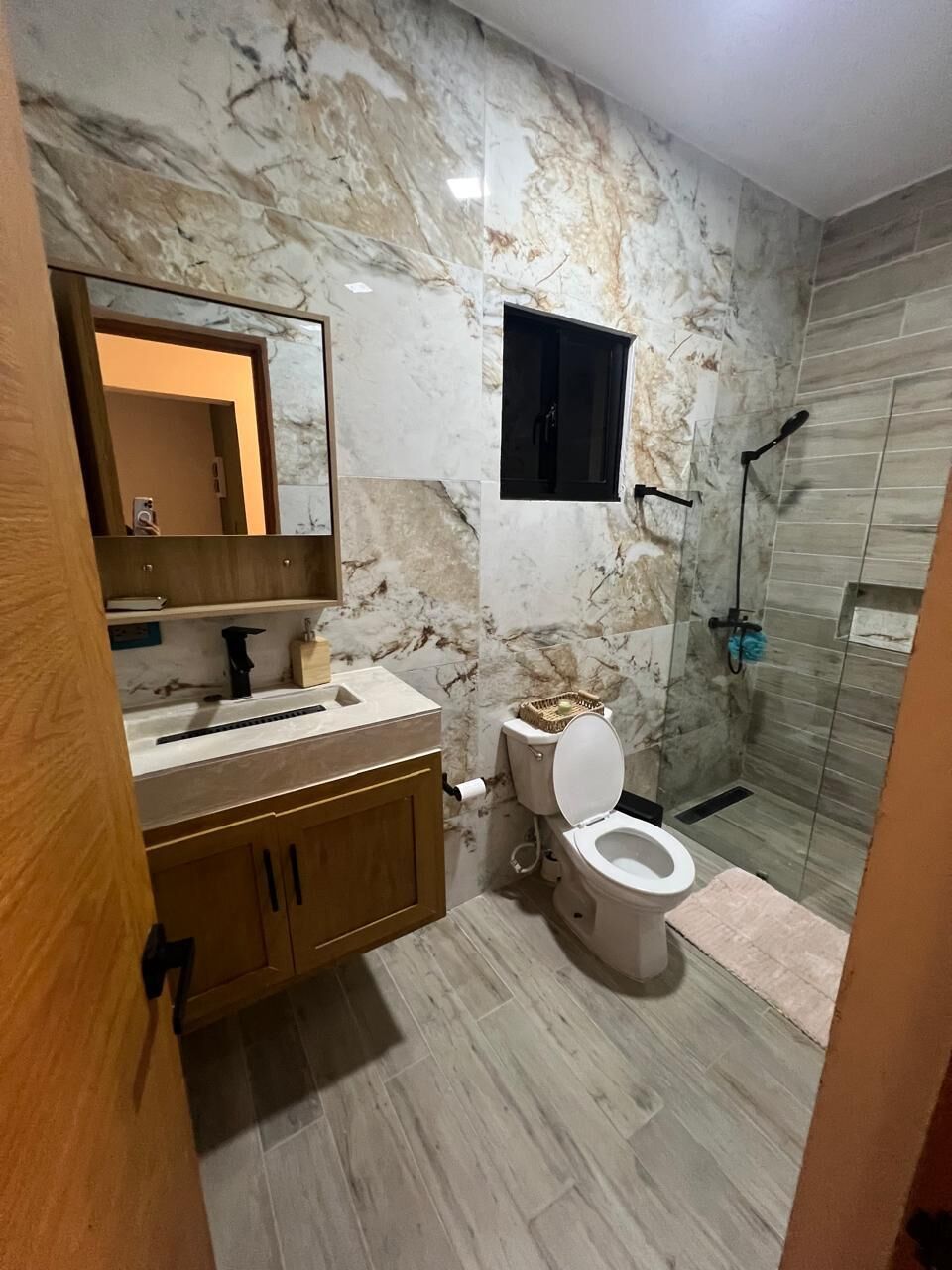 Baño con pared de mármol, tocador de madera, inodoro y zona de ducha.