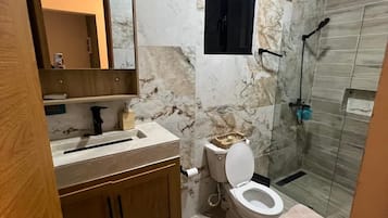 Baño con pared de mármol, tocador de madera, inodoro y zona de ducha.