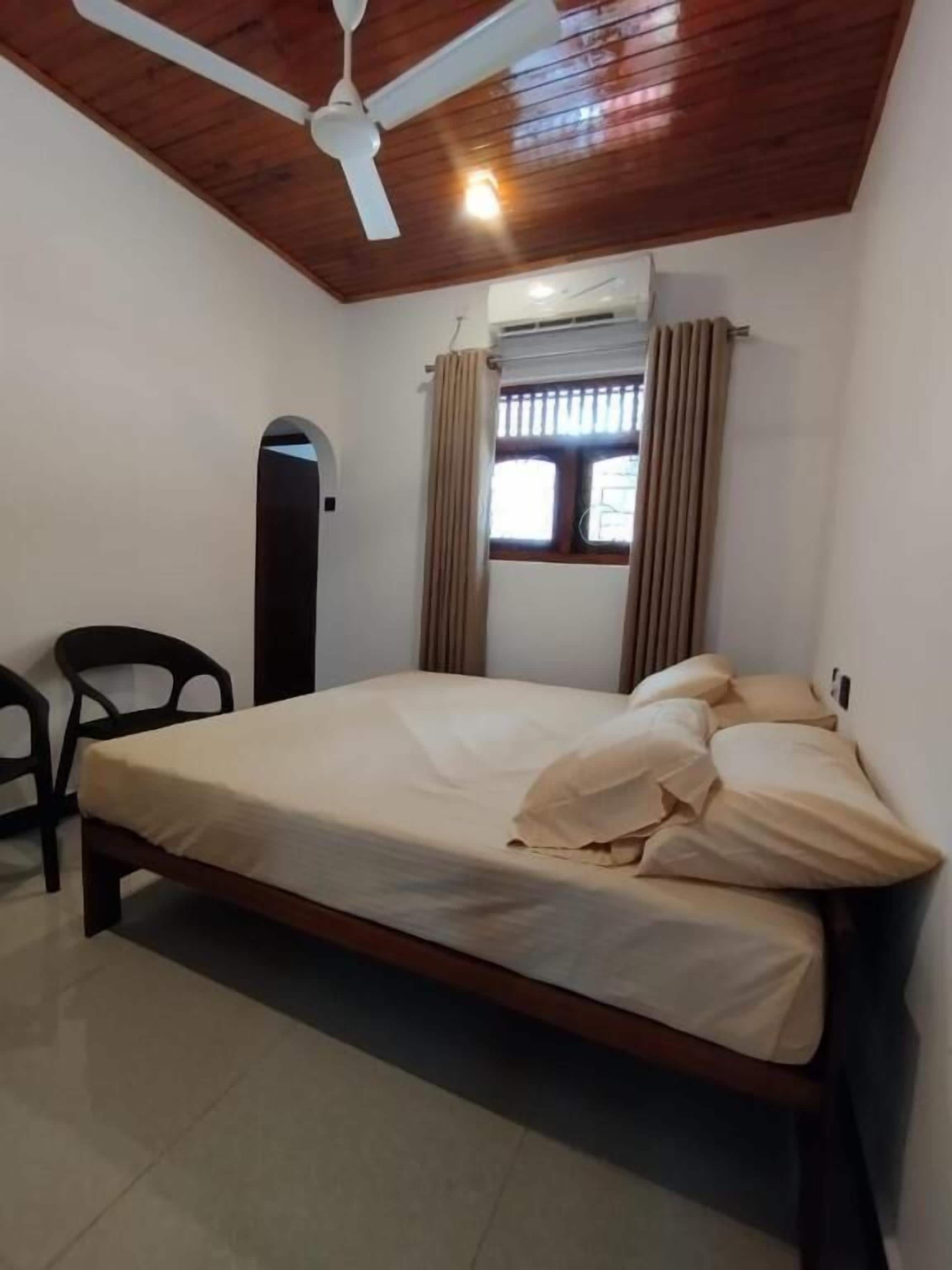Ein Schlafzimmer mit Bett, Deckenventilator, Fenster mit Vorhängen und einem Sessel.