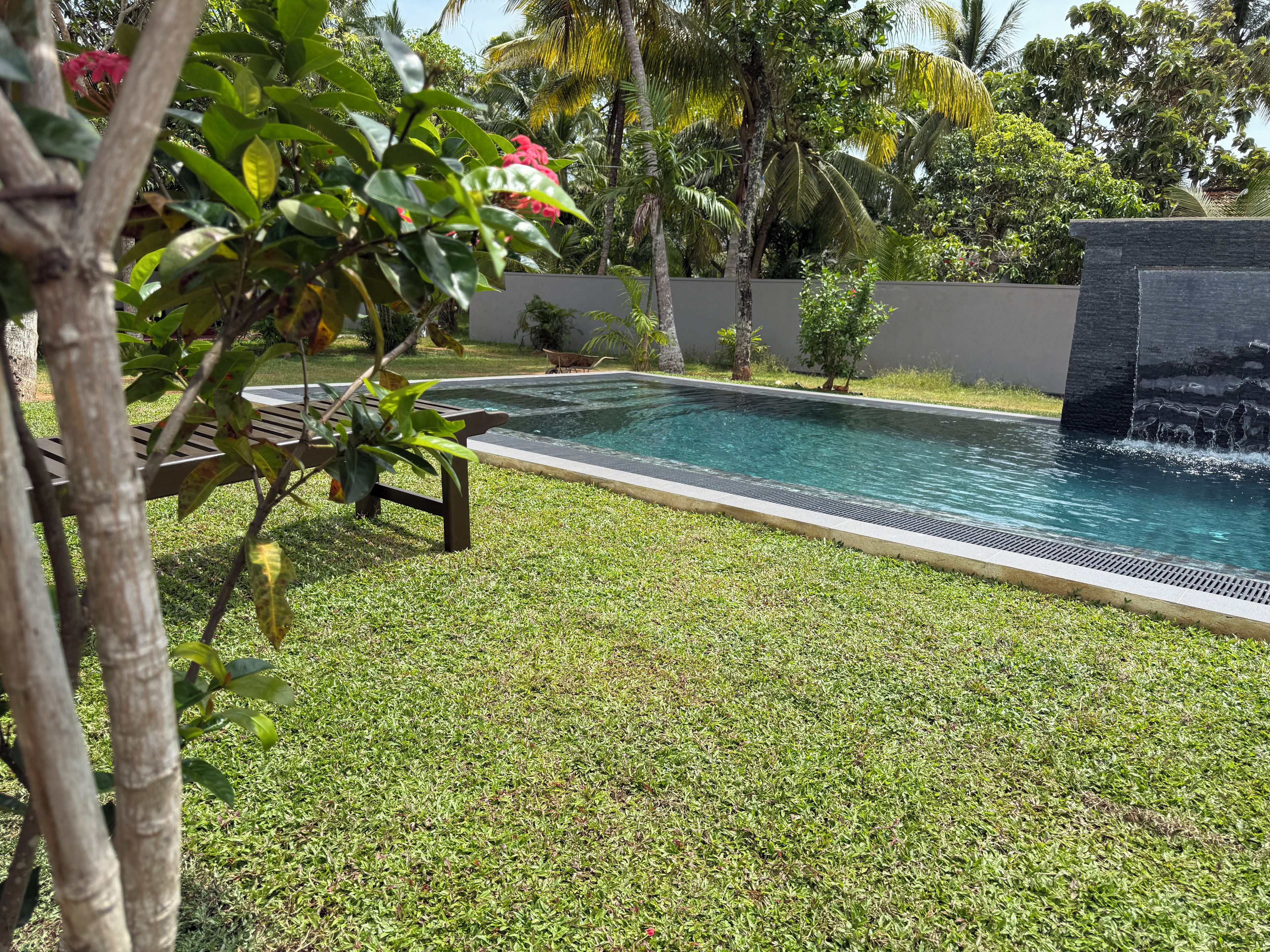 A rectangular swimming pool with a waterfall feature, surrounded by a well-maintained lawn and various tropical plants.