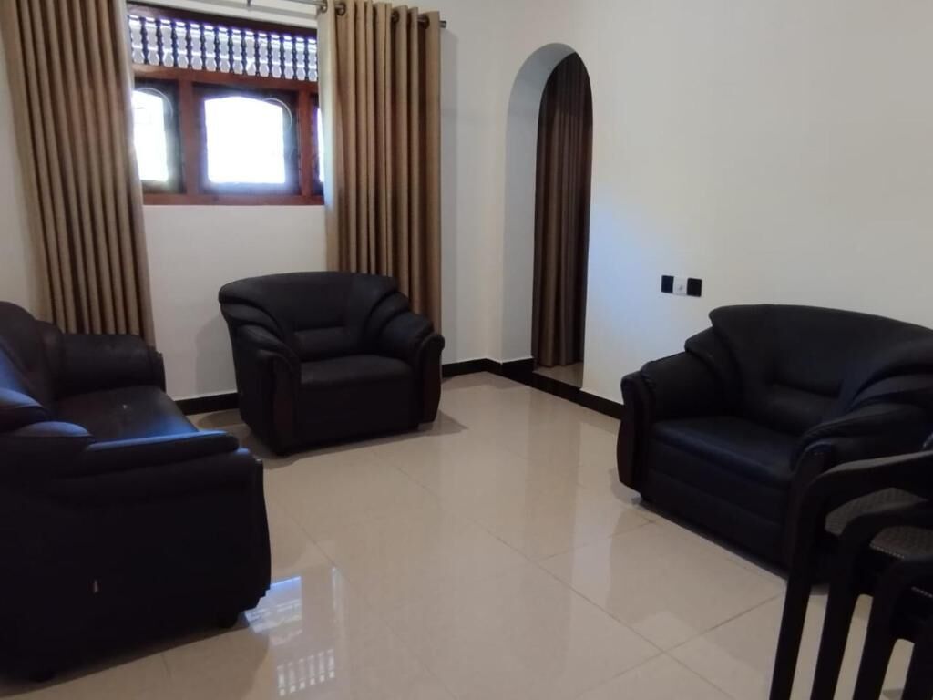 Un salón con tres sofás negros, una ventana con cortinas y suelo de baldosas.