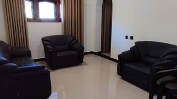 Un salón con tres sofás negros, una ventana con cortinas y suelo de baldosas.