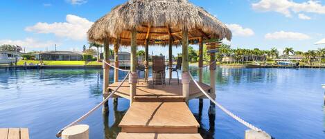 Un muelle de madera que lleva a un pabellón con techo de paja, con sillas y mesas, con vistas a aguas tranquilas.