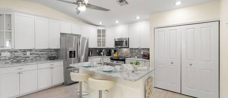 Una cocina moderna con armarios blancos, electrodomésticos de acero inoxidable, una encimera de mármol y un ventilador de techo.