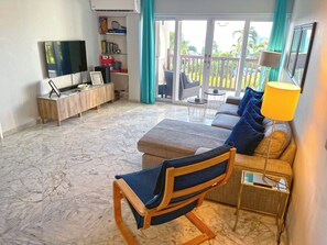 A living room with a sofa, a chair, a TV, and a bookshelf. - St Thomas 2BR Oceanside Villa with Views: Steps to Beach, Pool & Restaurants (Charlotte Amalie)