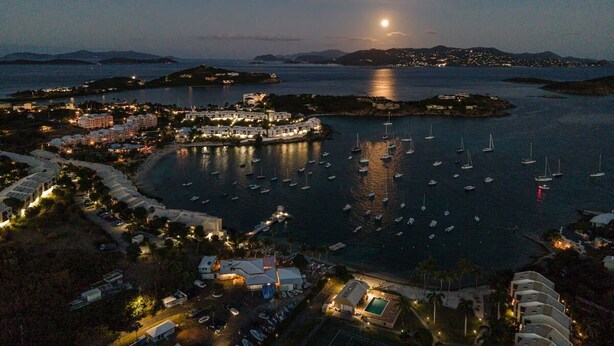 Uma cidade costeira à noite com um porto repleto de barcos, um edifício iluminado em destaque e uma lua crescente no céu.
