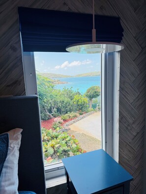 A room with a large window offering a view of a coastal landscape, a blue table, and a chair.