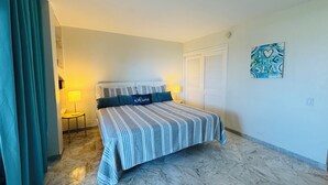 Quarto de hotel com uma cama, duas luminárias de cabeceira, uma porta, uma obra de arte na parede e piso de mármore.