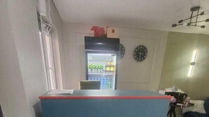 Un espacio habitable compacto con kitchenette, un refrigerador lleno de bebidas, un reloj en la pared y una pequeña mesa con una silla.
