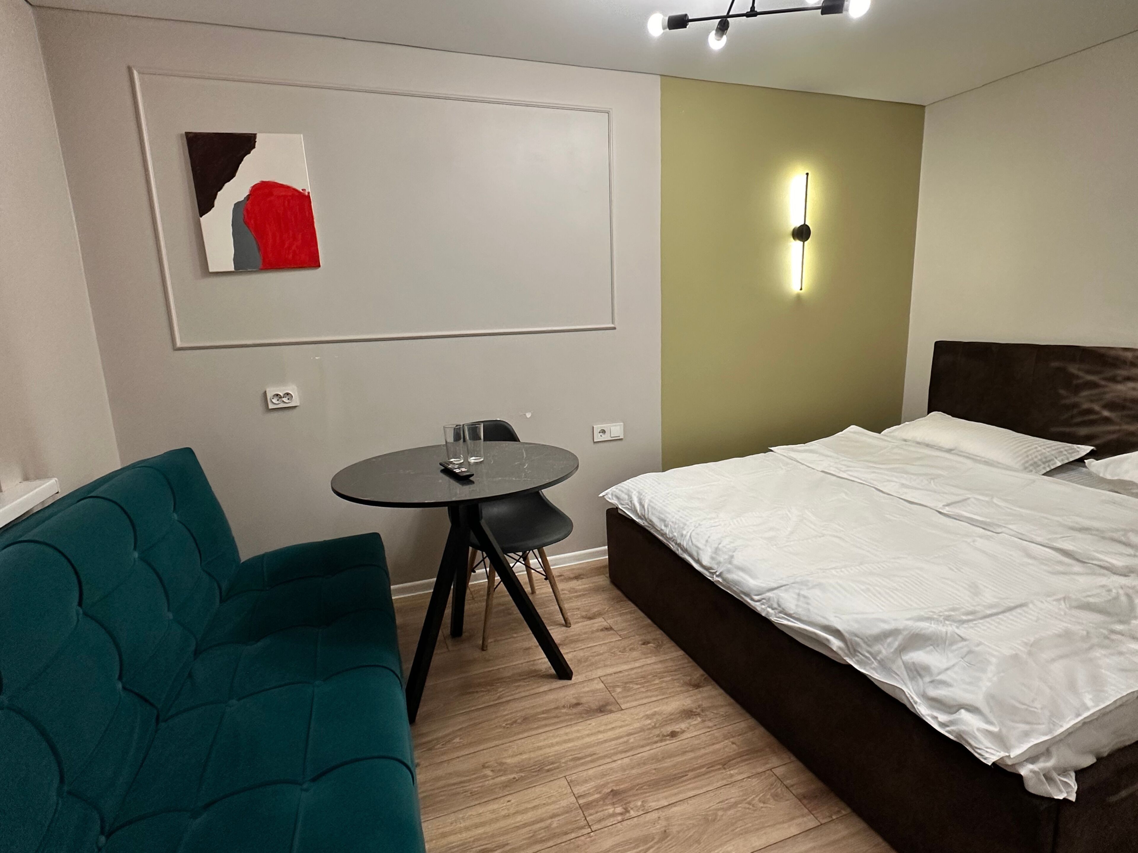 Una habitación compacta con cama, sofá verde, una mesita redonda con dos vasos y una lámpara de pared.