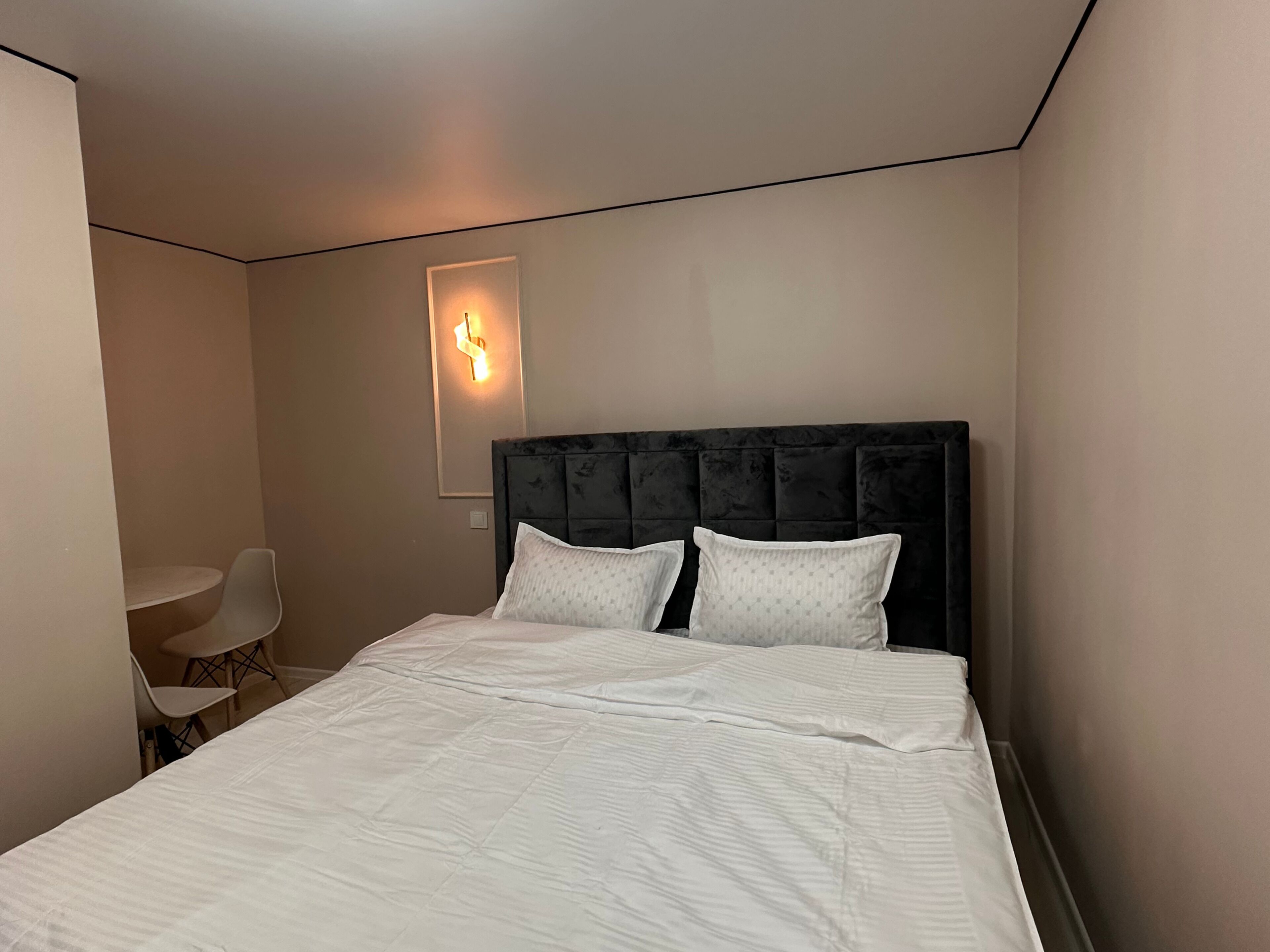 Een netjes opgemaakt bed met een donkere hoofdbord en twee kussens, een kleine tafel met een stoel, en een wandlamp.